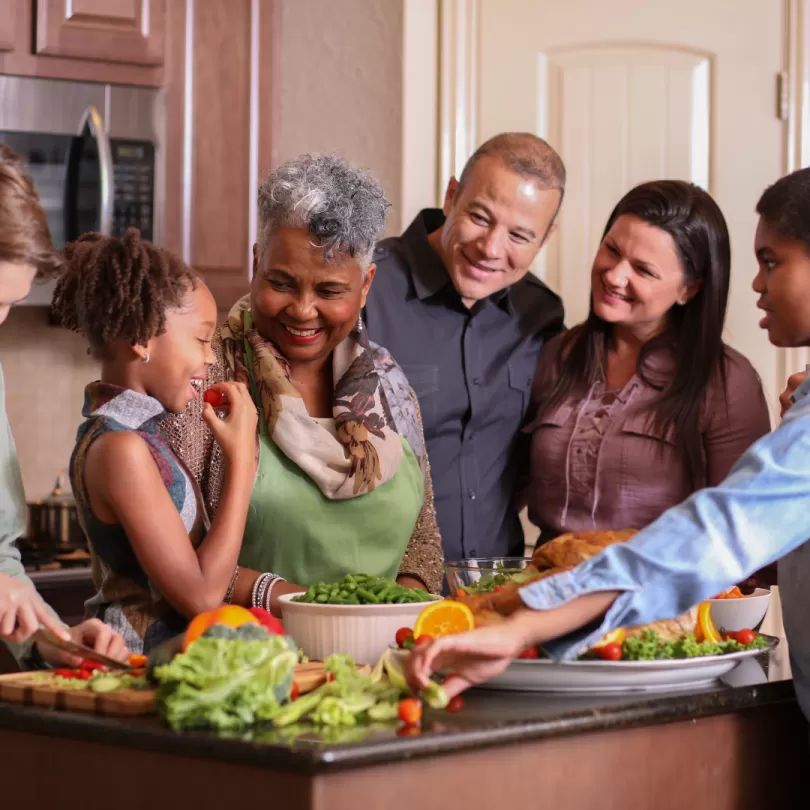 A family gathers in the kitchen as they cook a healthy holiday meal. A little girl stands at the counter and eats a cherry tomato.