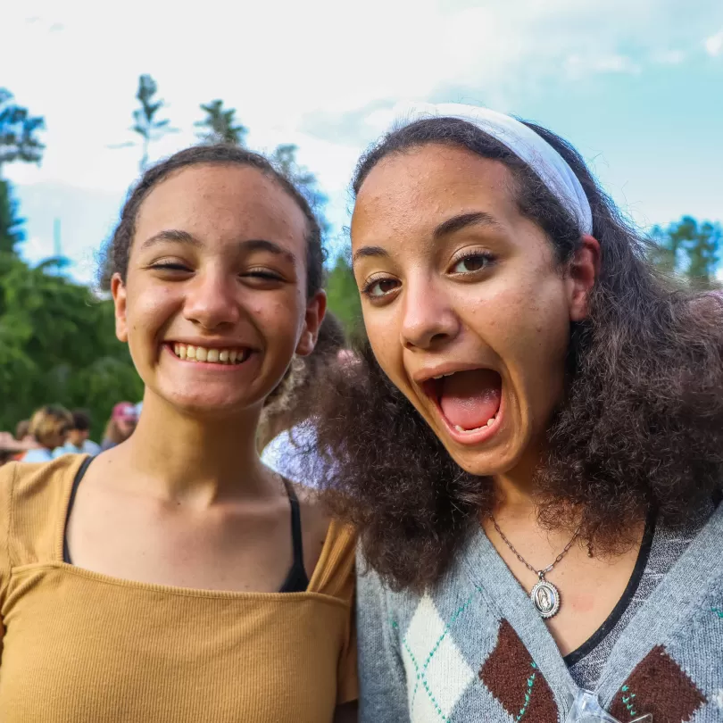 two girls who appear related with a big smile and a funny face in the camera with a group of kids in the background