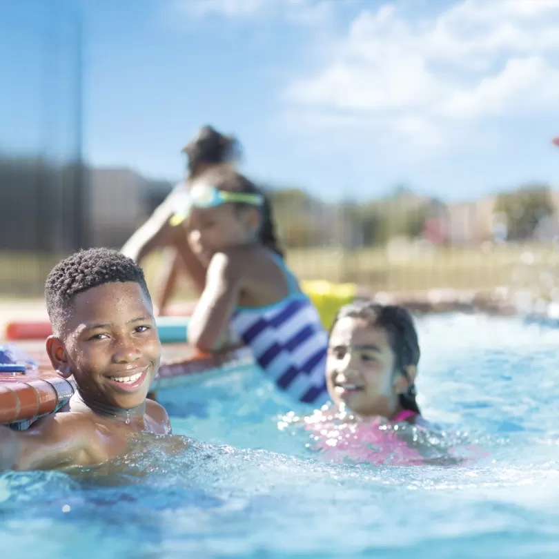 A group of kids in the pool smiling and having fun, boy is close and looking into camera with a big smile. Lifeguard in the background