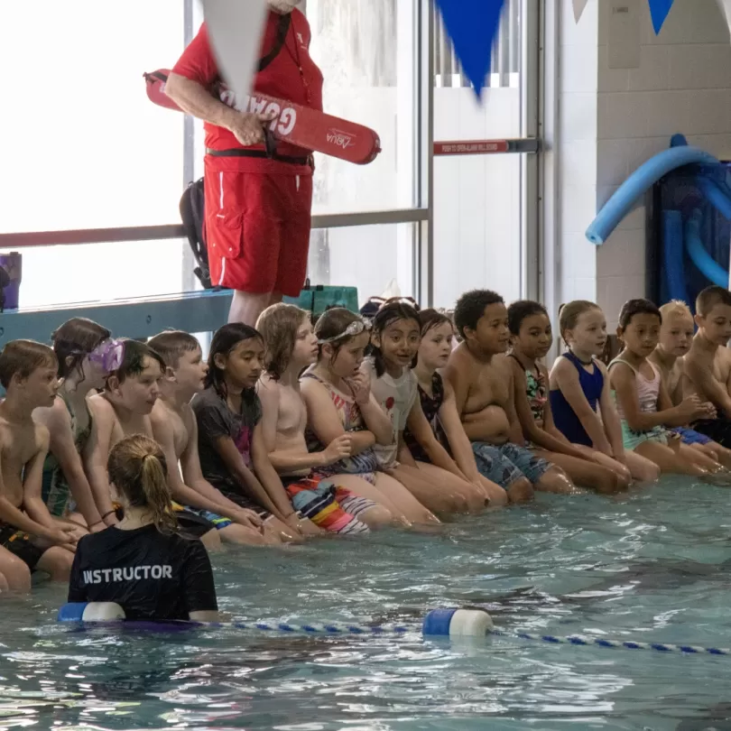 A large group of kids sit on the side of a pool listening to instructor. There is a lifeguard standing in the background.