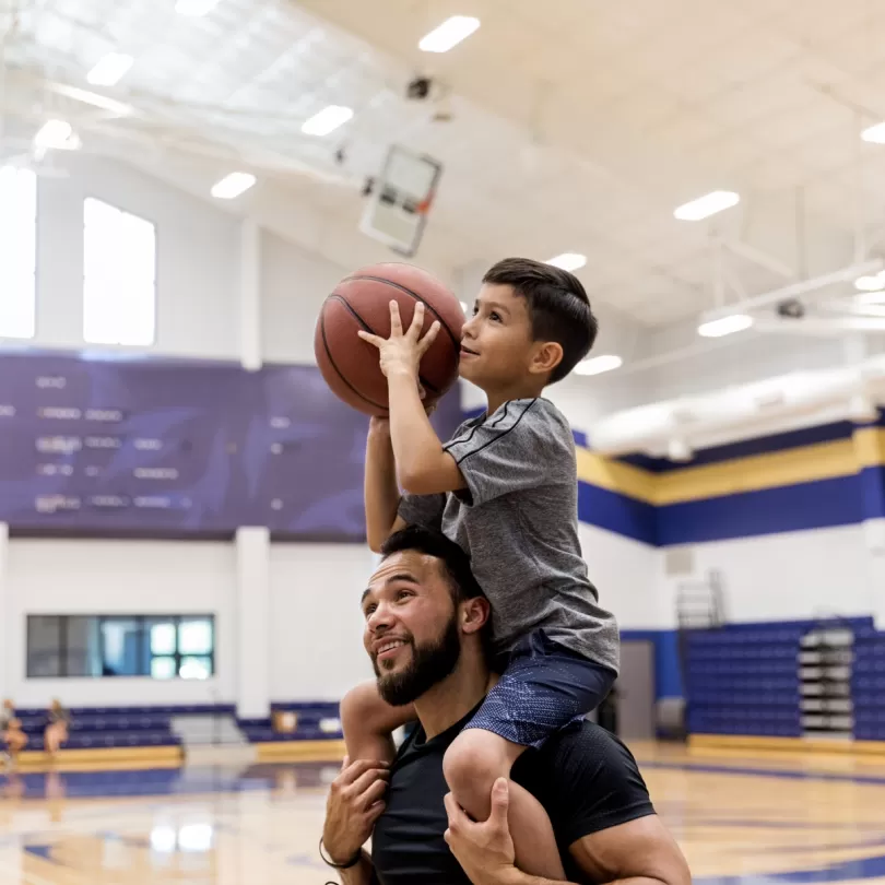 Father holding son on shoulders, son has basketball in hand and is in shooting motion