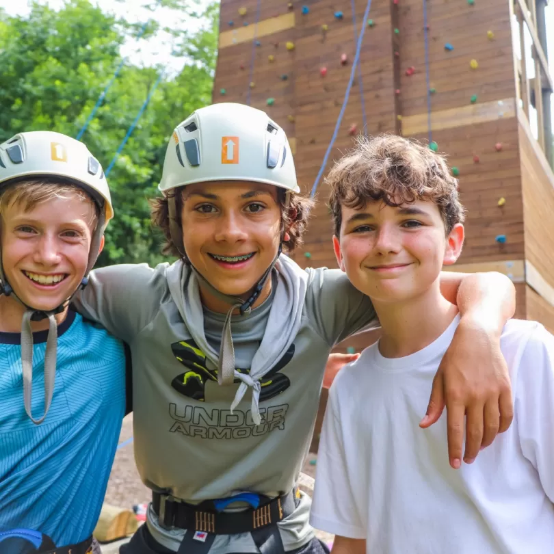 Three boys, two wearing helmets and ready to climb on the climbing wall in the background. All three kids have a big smile. 