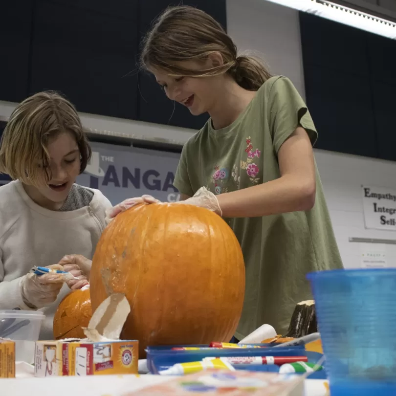 Two pre-teen girls work together to carve a pumpkin, both are laughing.