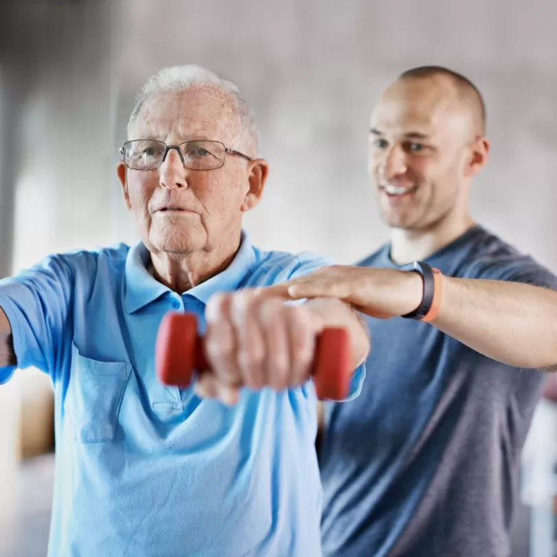Older man lifting weights at the director of a trainer who is standing behind him helping guide his arm.