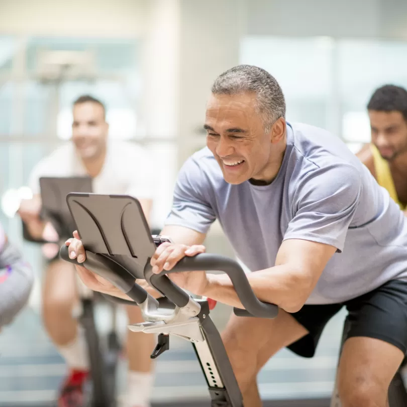 Middle age man on cycle machine with big smile along with a large group of diverse people in the background also cycling 