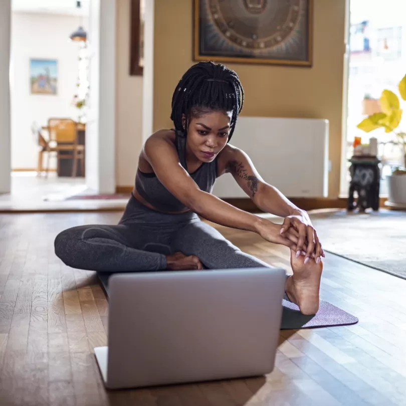 woman sitting on floor in what appears to be a living room doing a leg stretch while looking at a laptop screen