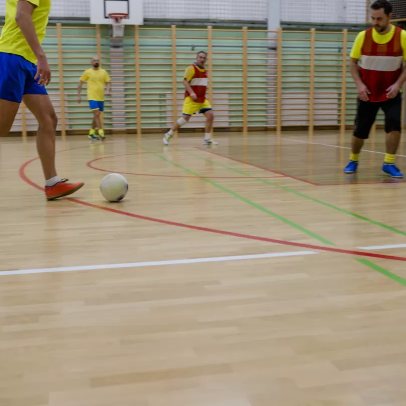 People kicking a soccer ball around inside a gym wearing different colored jerseys.