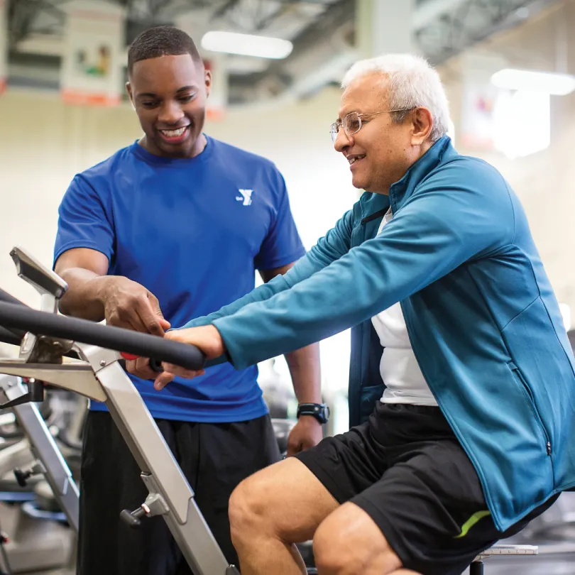 A personal trainer helps a man use a stationary bike at the gym.