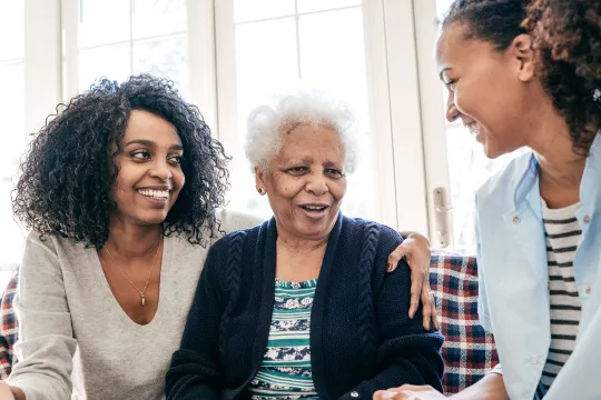 A grandmother sits on the couch with her two grown granddaughters sitting next to her on either side. One granddaught has her arm around her grandmothers' shoulder. They are all smiling and talking with each other.