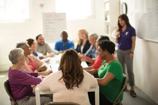 Group of people sit at a table looking at a booklet and paying attention to a teacher who is talking near a whiteboard