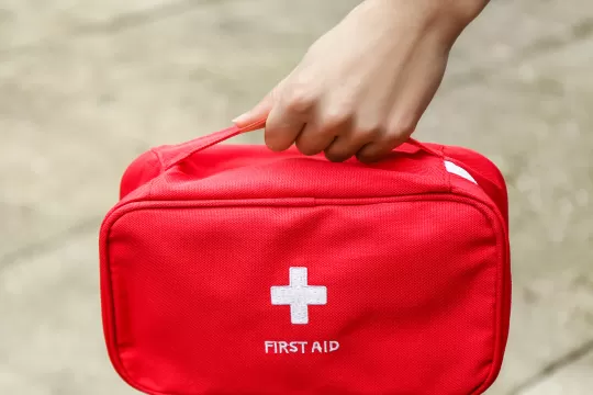 A hand carries a red bag that says "First Aid" below a white cross symbol.