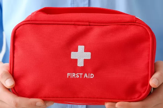 Two hands hold a red bag that says "First Aid" below a white cross symbol.