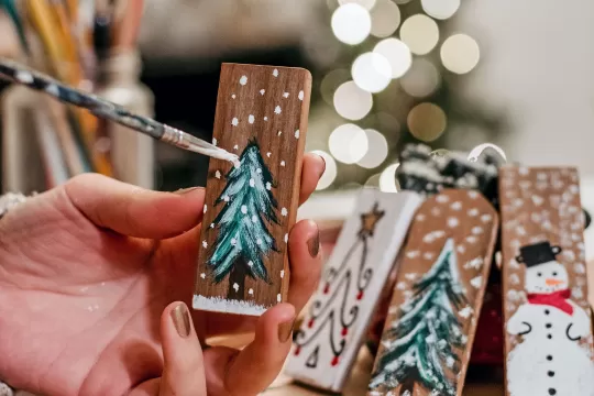 A closeup of a pair of hands painting a Christmas tree on a piece of wood. There are other holiday pieces in the background.