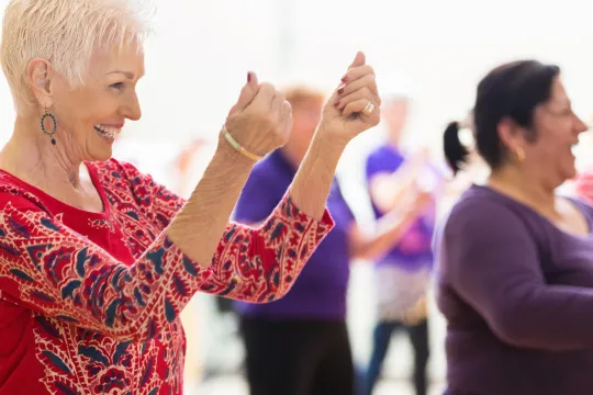 An older woman smiles as she line dances with others in a dance class.