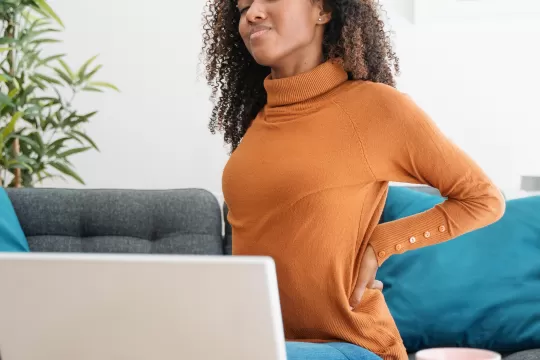 A woman sits on a couch in front of a computer. She is holding her lower back.