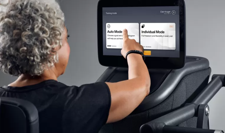An older woman uses touch screen settings on gym equipment.