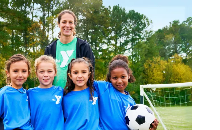 A volunteer sports coach stands outside next to the youth soccer team on the field. They are all smiling at the camera.