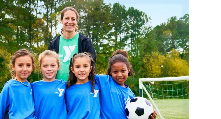 A volunteer sports coach stands outside next to the youth soccer team on the field. They are all smiling at the camera.