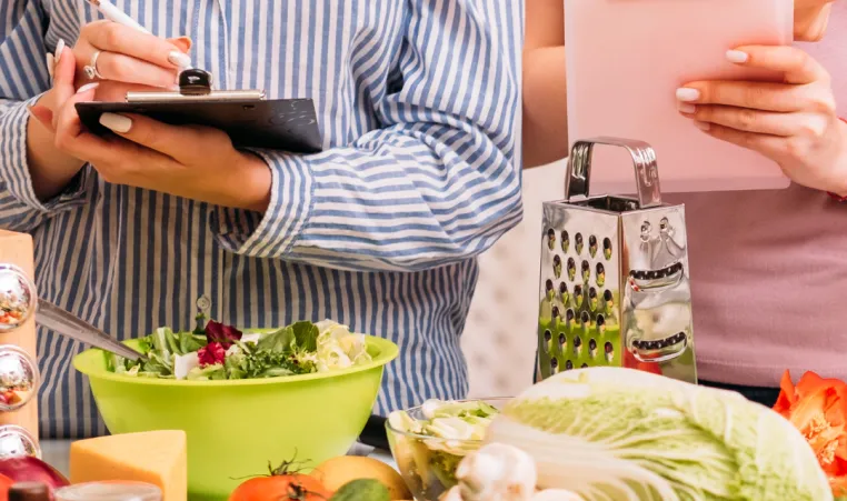 Two people stand in ftont of a counter covered in vegetables. Both people are holding clipboards and writing down information.