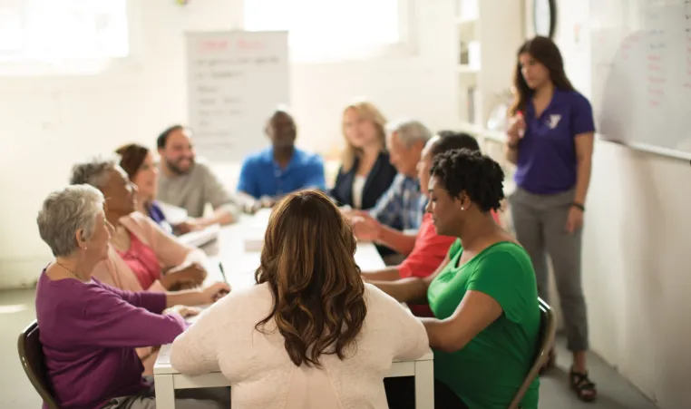 Group of people sit at a table looking at a booklet and paying attention to a teacher who is talking near a whiteboard