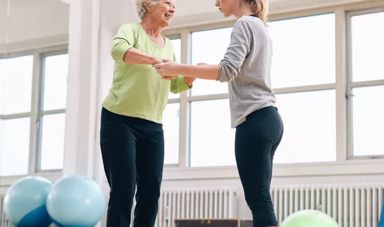 An older woman balancing on a balance ball. Another woman supports her by holding her hands.