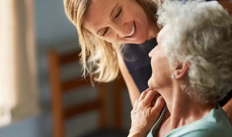 Elderly woman with caregiver. They are smiling at each other.
