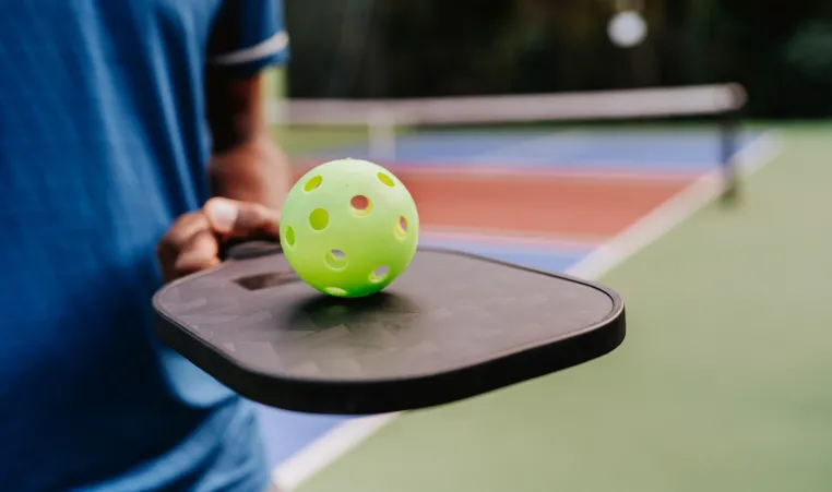 A closeup of someone holding a pickleball paddle and pickleball while on a court.