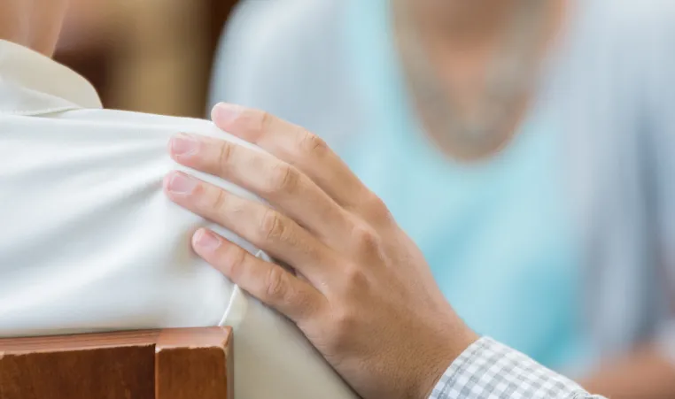 A person places their hand on someone else's shoulder in support during a grief support group.