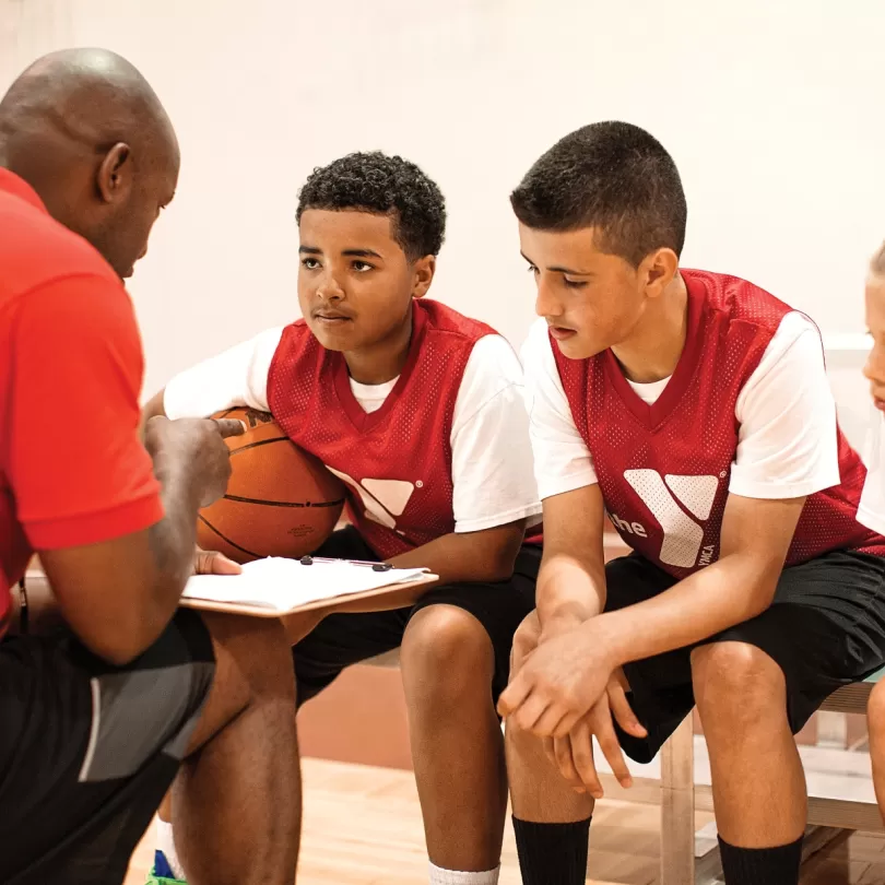 Three youth basketball players wearing Y jerseys sit on a bench while the coach lays out a plan on a clipboard